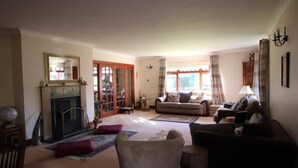 A living room with a fireplace, sofa, and armchairs. There is a large window with curtains, a chandelier, and a mirror on the wall.