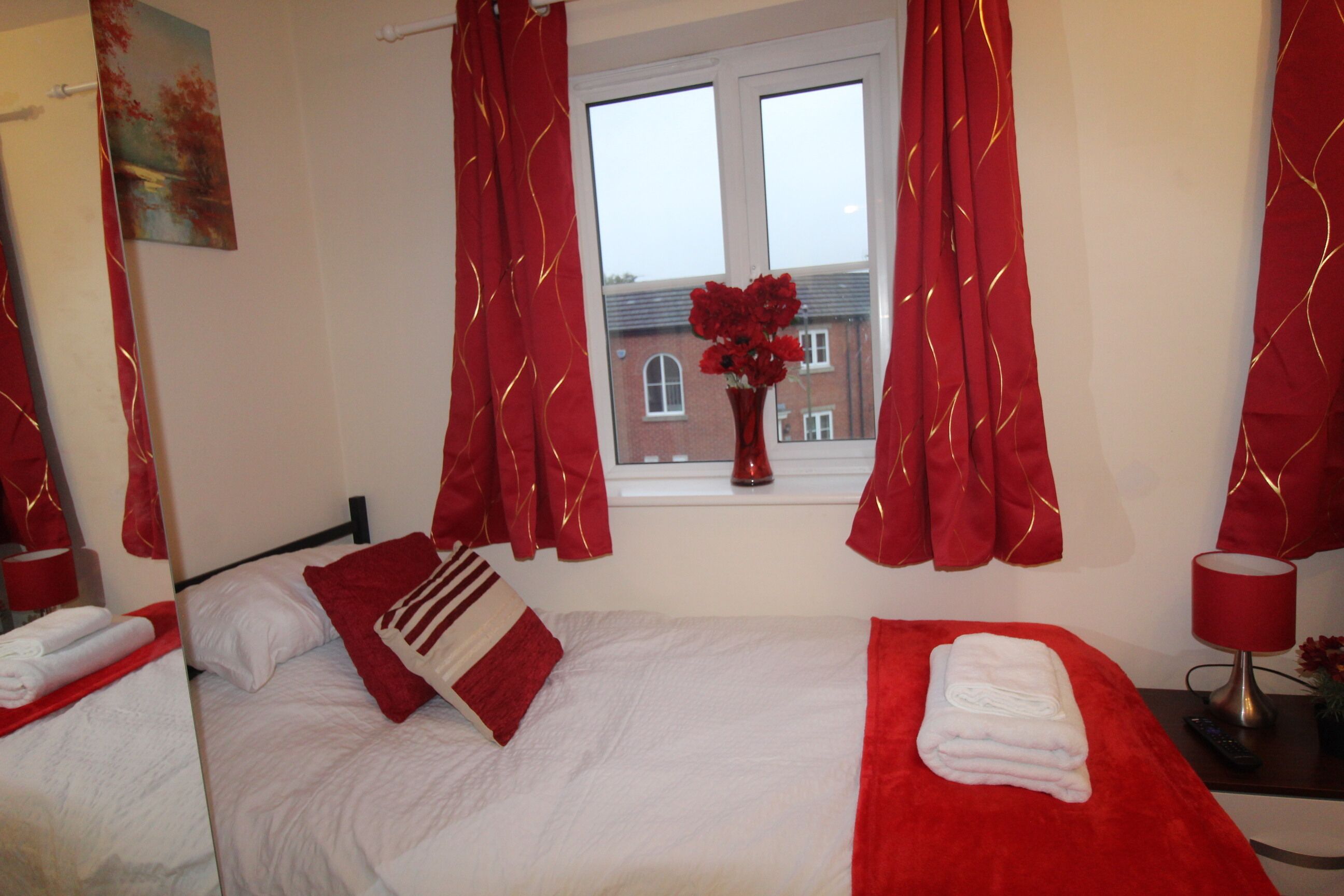 A hotel room with a bed, a window with red curtains, a vase with flowers, and a lamp on a bedside table.