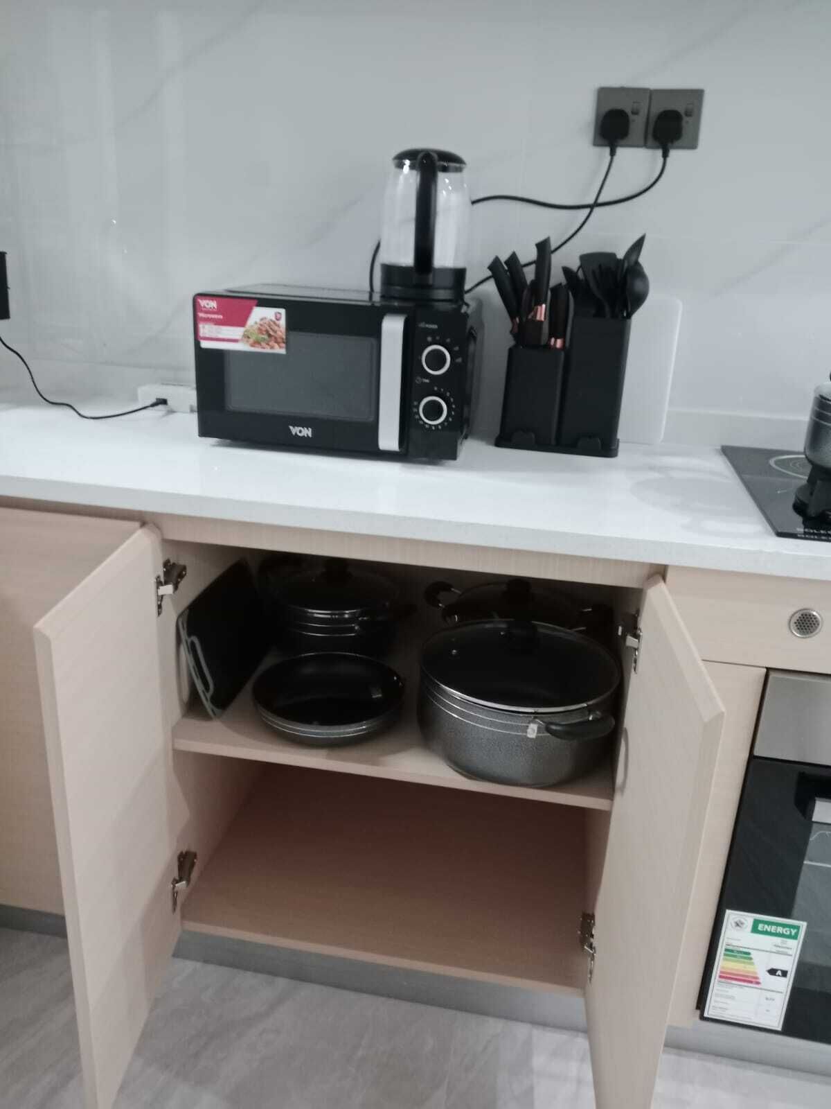 Una cocina con microondas, cafetera y un juego de cuchillos sobre la encimera. Hay ollas y sartenes guardadas en un armario debajo.
