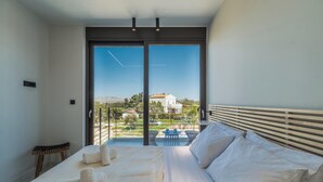 A modern bedroom with a large bed, a small table, and a view of the outdoors through a sliding door. - Nos Aqua Villas (Chania)