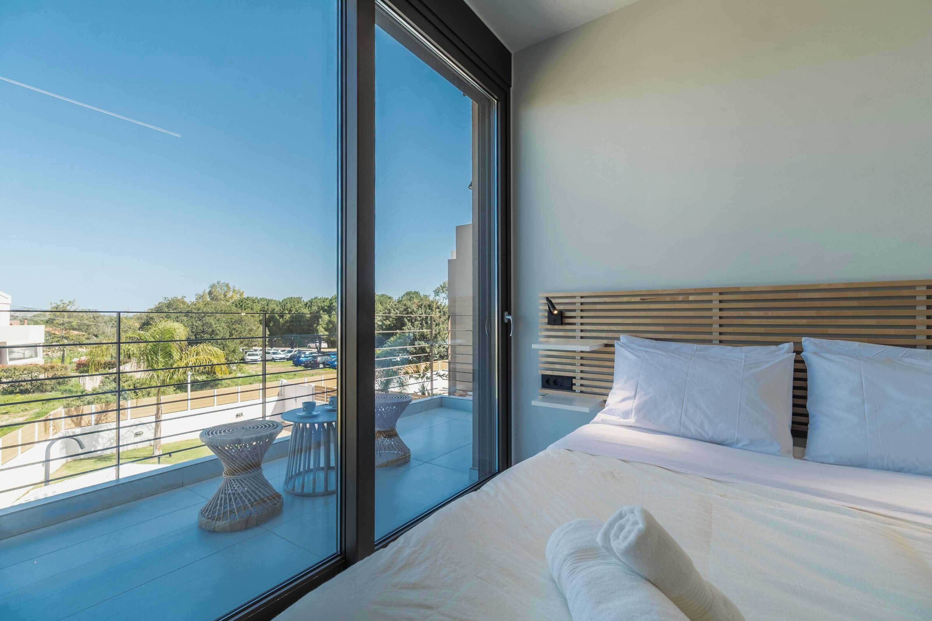 A modern bedroom with a large window, a bed with white bedding, and a view of the cityscape.