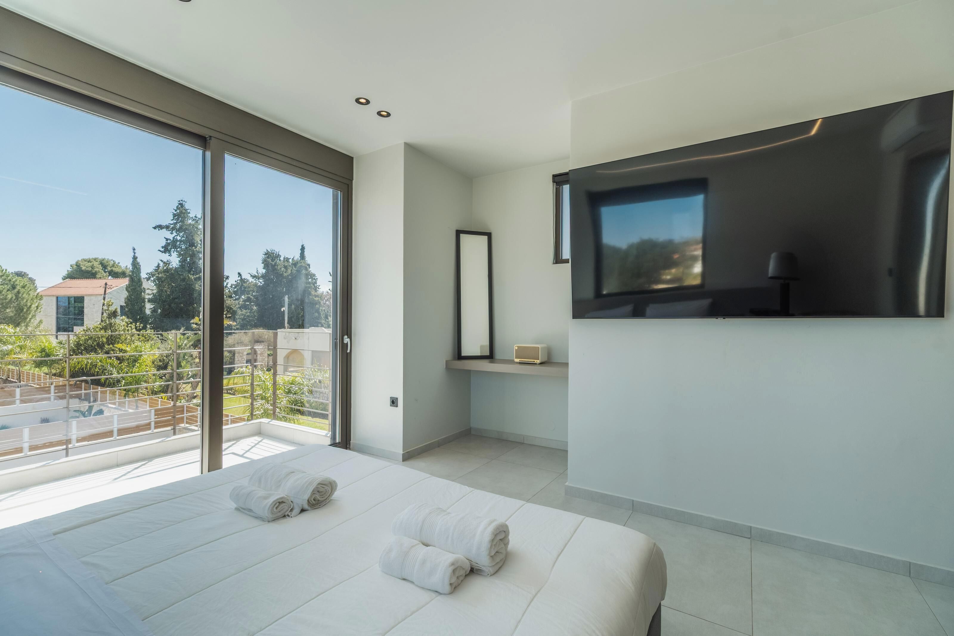 A modern hotel room with a large bed, a TV mounted on the wall, and a balcony with a view of the surrounding area.