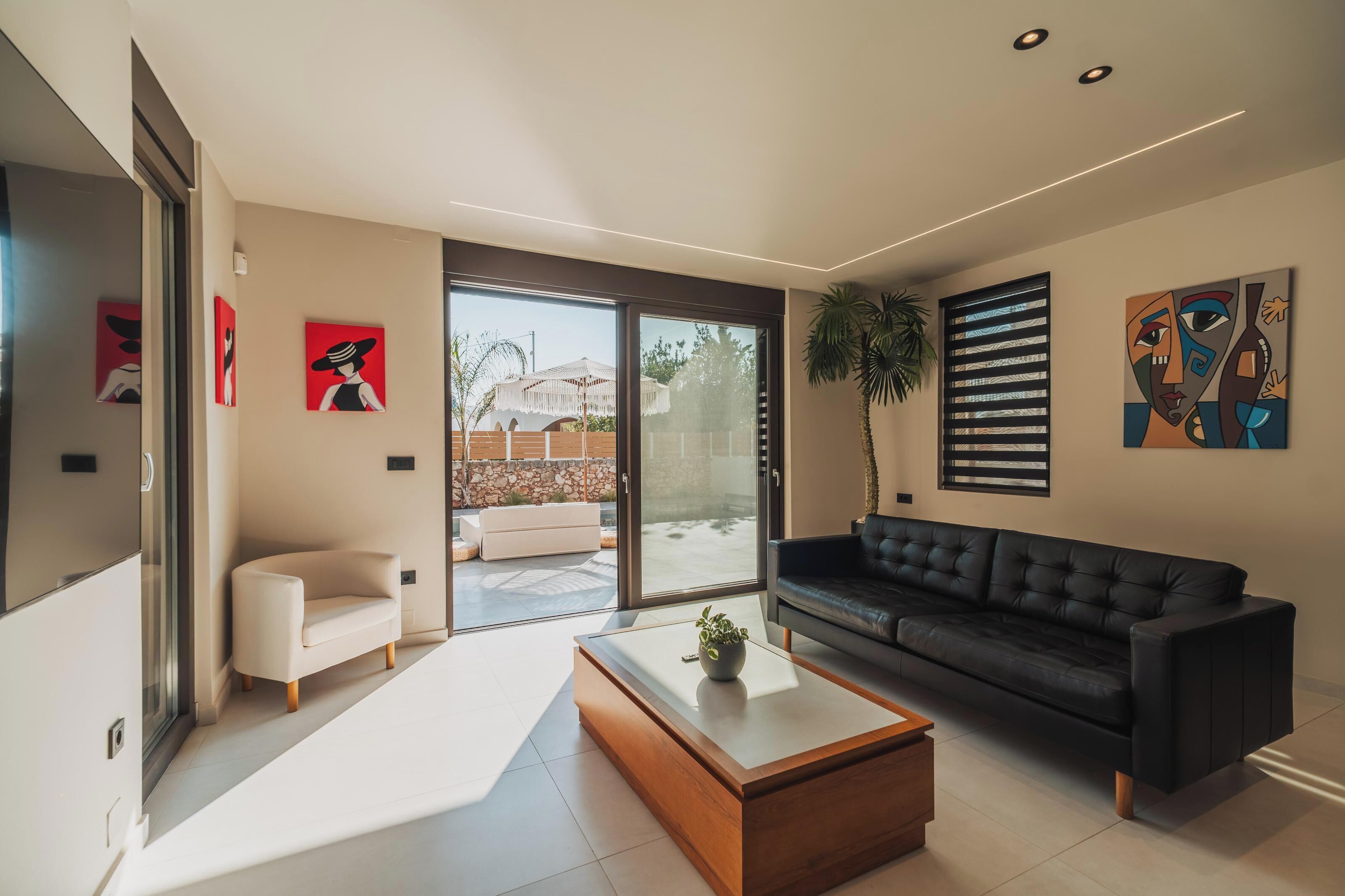 A modern living room with a black leather sofa, a white armchair, and a wooden coffee table. There are large windows and sliding glass doors leading to an outdoor patio.