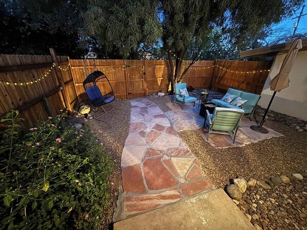 Une terrasse à l’arrière de la maison, avec un sol en pierre, une clôture en bois et des guirlandes lumineuses.