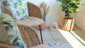 A wicker chair with a cushion, a potted plant, and a colorful container on a tiled floor.