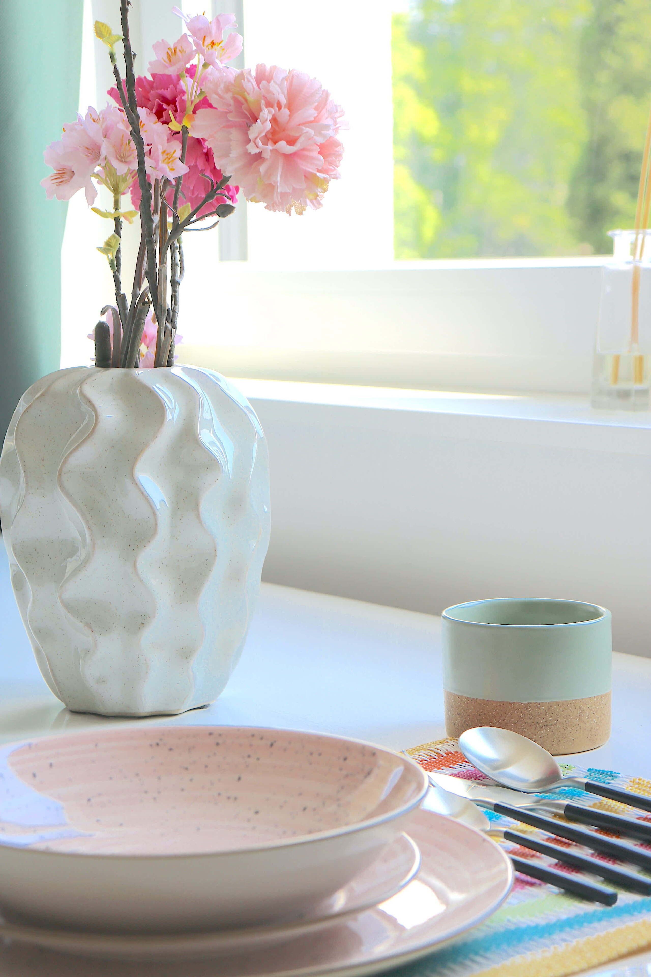 Un jarrón con flores, una taza y una cuchara sobre una mesa cerca de una ventana.