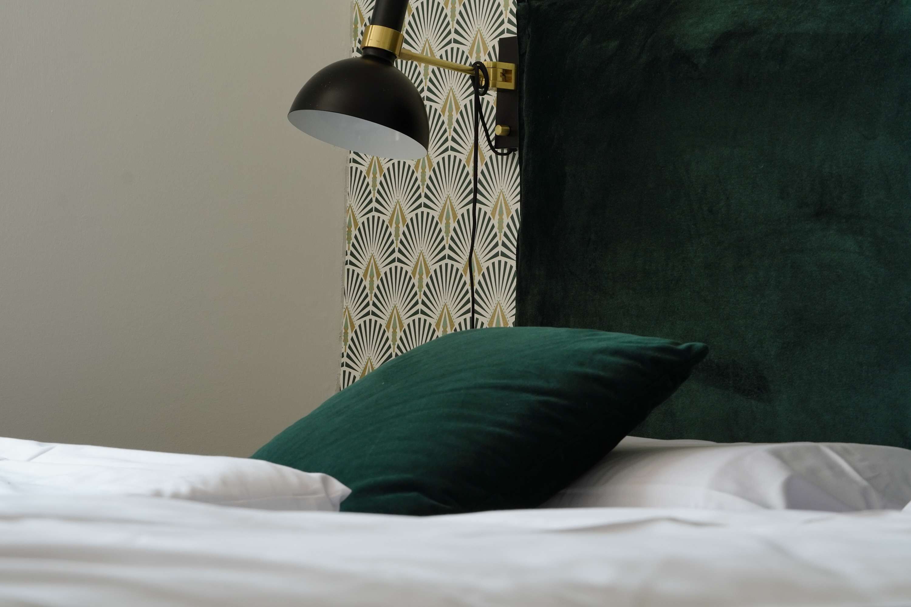 Cama com roupa de cama branca e almofada verde, luminária de mesa preta e papel de parede estampado.