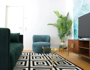 Uma sala de estar com sofá, uma poltrona, uma mesinha, planta em vaso, televisão e tapete estampado.