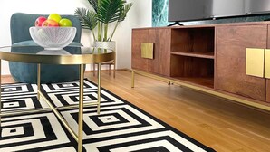 A living room with a round glass table, a blue armchair, a wooden cabinet, a large TV, and a patterned rug. - 1 Zimmer Apartment mit Blick auf die Stadt (Gmunden)