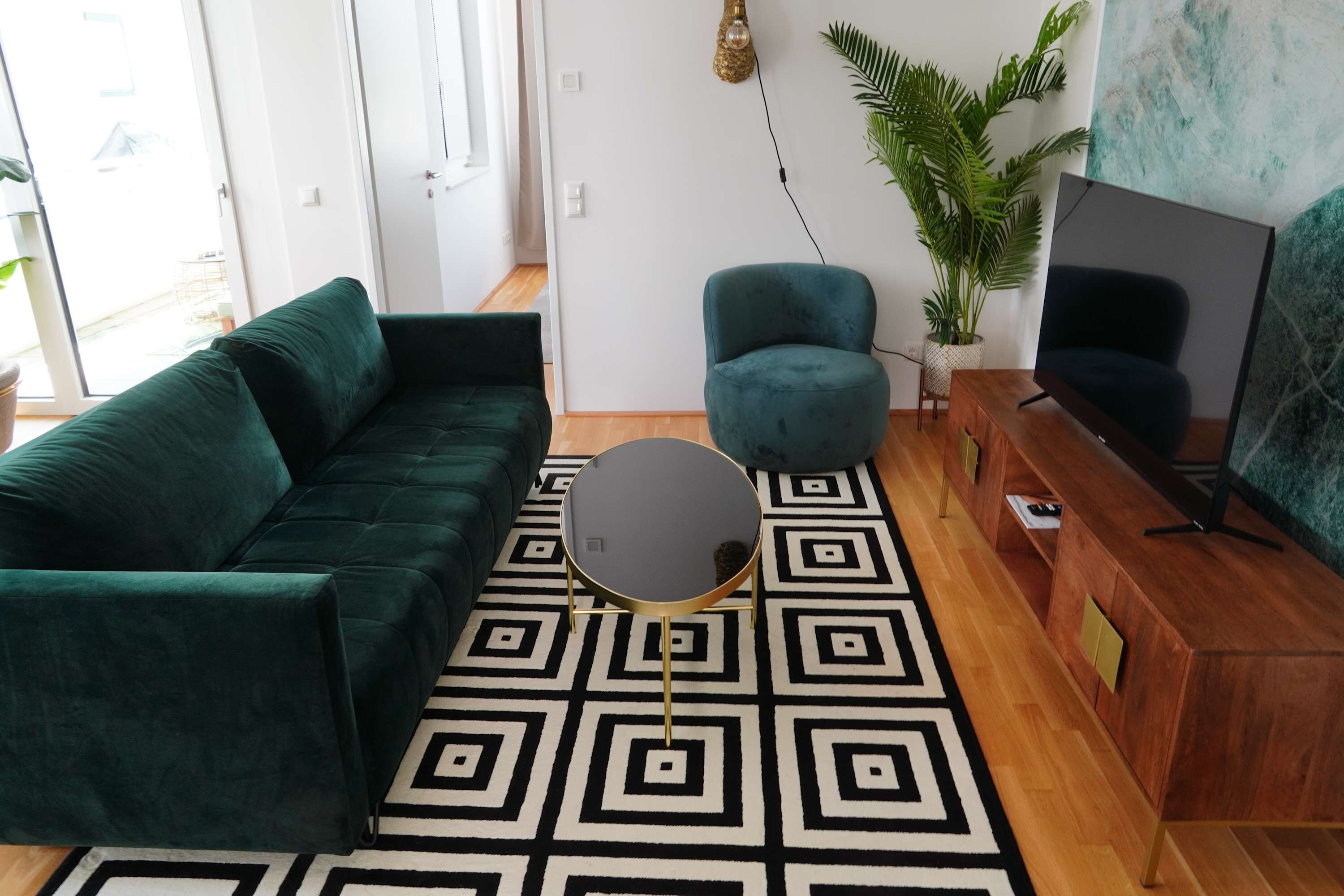 A living room with a dark green sofa, a round coffee table, a teal armchair, a wooden TV stand, and a patterned rug.