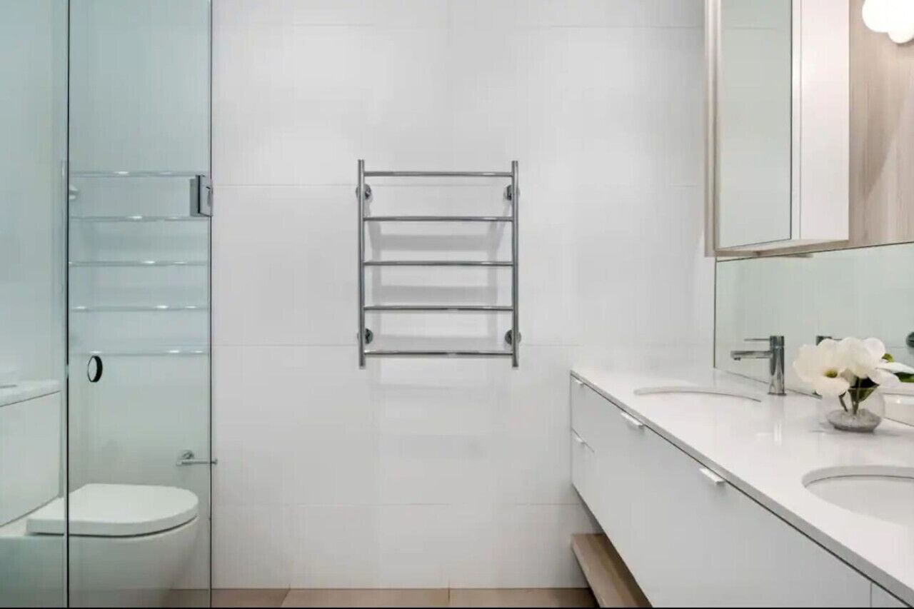 A modern bathroom with a glass shower enclosure, a white toilet, a metal towel rack, and a vanity with a mirror and a vase of white flowers.