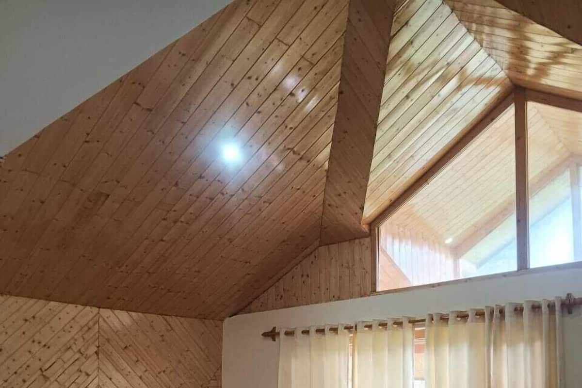 Une pièce avec un plafond en bois, un lit doté d’une tête de lit à motifs, une fenêtre avec des rideaux et une petite table entourée de chaises.