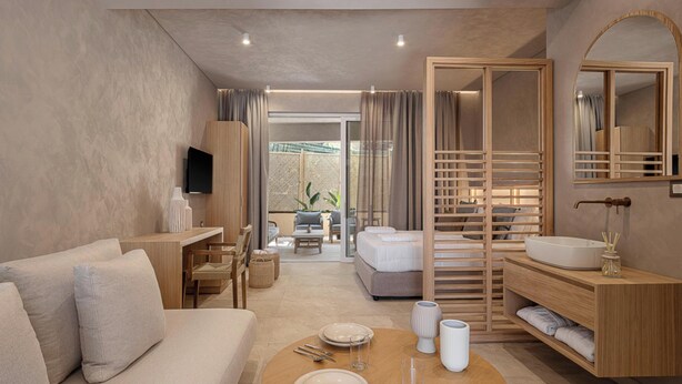 Ein modernes Hotelzimmer mit einer Couch, einem Esstisch gedeckt mit Tellern und Besteck, einem hölzernen Schminktisch mit Spiegel und einem Bett mit Holz Kopfteil.