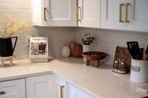 Eine Küche mit weißen Schränken, einem kleinen Holzschemel, einer Vase mit getrockneten Blumen, einem dekorativen Schild und einer Holzüble auf der Arbeitsfläche.