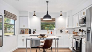 Cocina moderna con armarios blancos, electrodomésticos de acero inoxidable, una isla central con taburetes, iluminación colgante y vistas a zonas verdes.