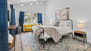 Ein Schlafzimmer mit einem Bett, einem gelben Sofa, einem gemusterten Teppich und einem Fenster mit Vorhängen.
