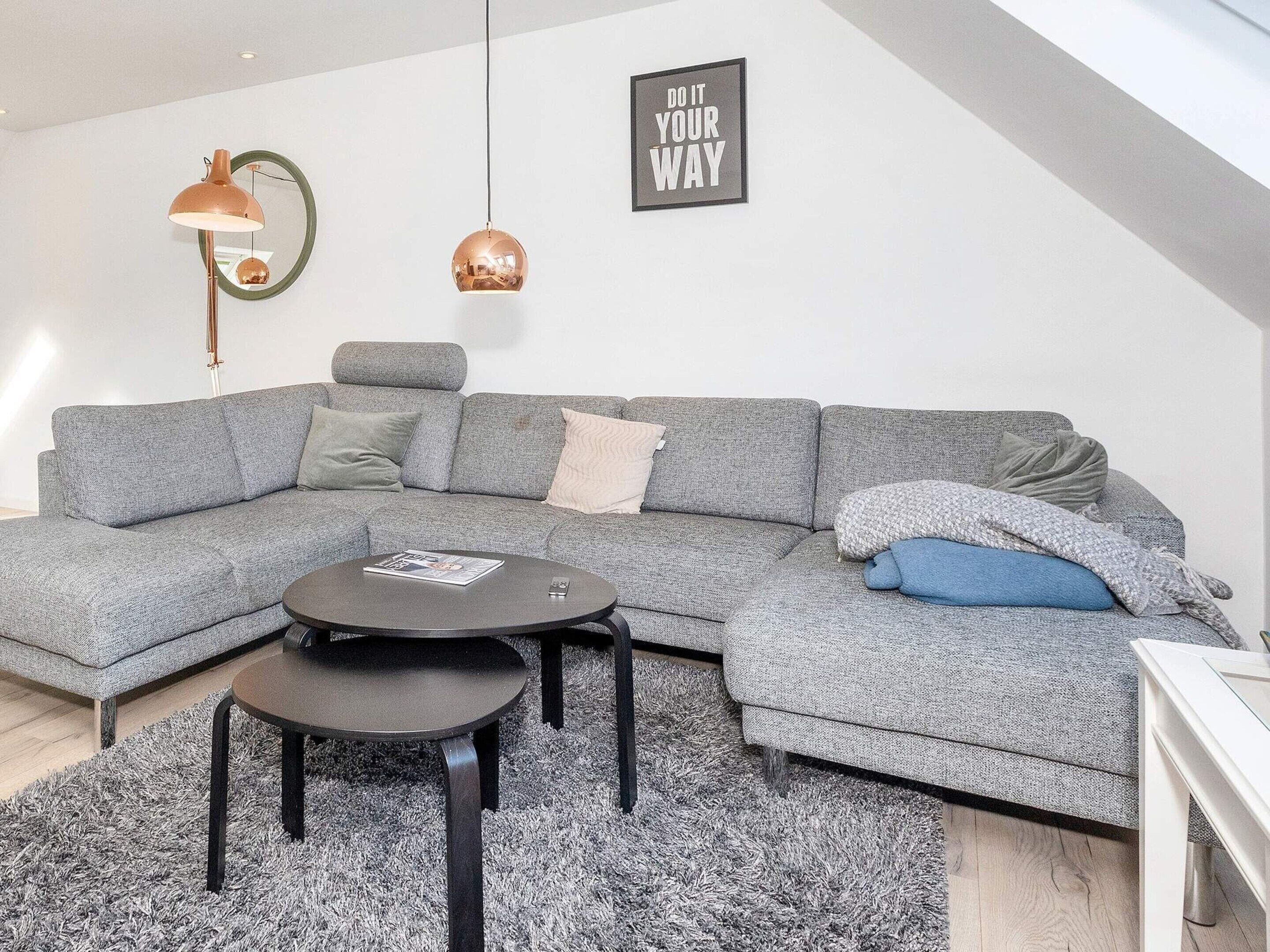 Een moderne woonkamer met een grijze modulaire bank, een ronde salontafel en een bijzettafeltje. Op de muur hangt een lijst met een motiverende quote en boven de bank hangt een hanglamp.
