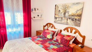 Een slaapkamer met een bed, een houten nachtkastje, een nachttafel, een wandplank en een schilderij.