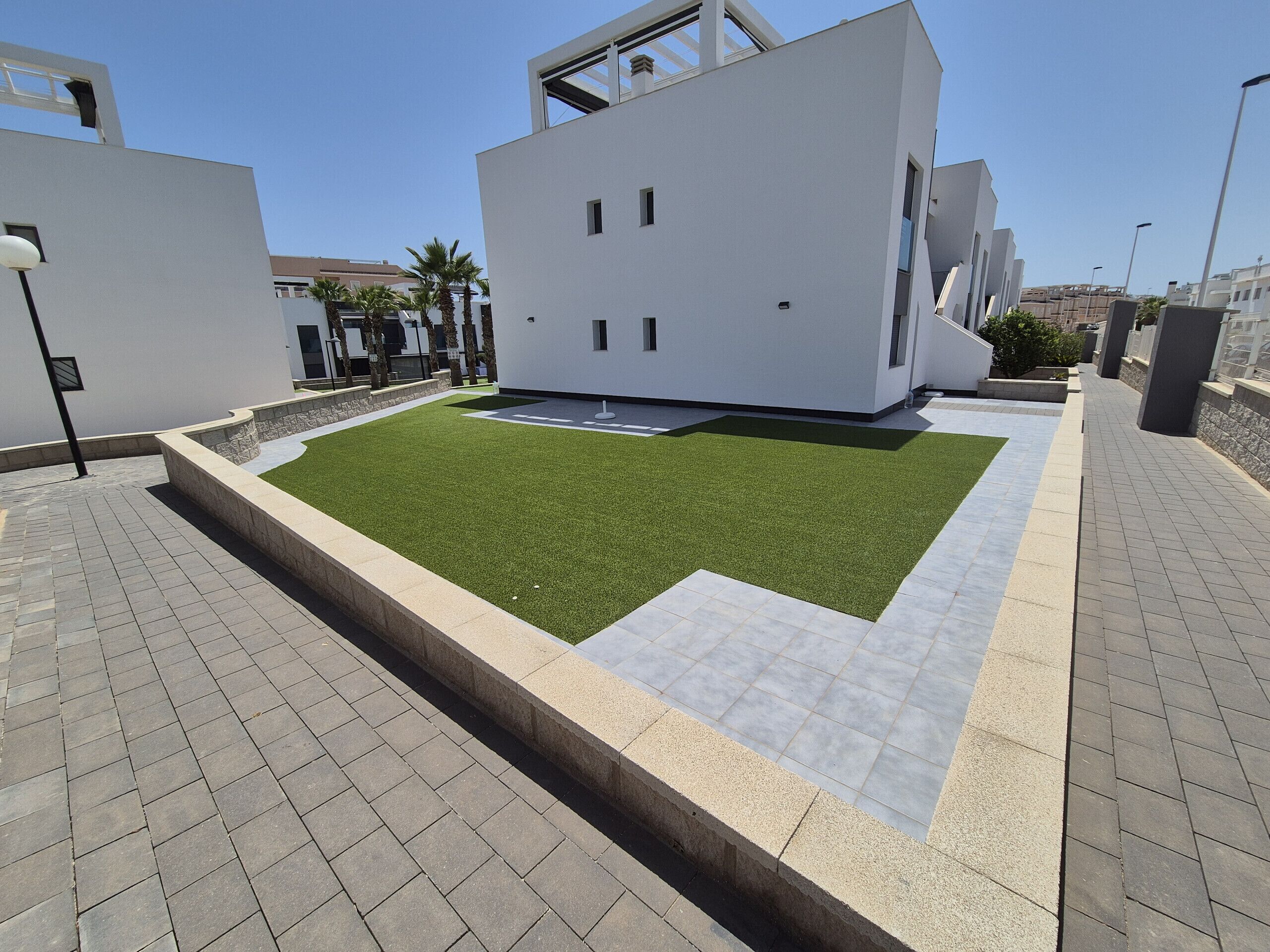 Un edificio moderno con techo plano, un patio rectangular con césped artificial y un camino pavimentado.