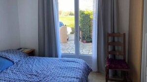 A bedroom with a bed, a chair, a window with curtains, and a view of a car and greenery outside. - 3-bedroom house for 6 people (Argelès-sur-Mer)