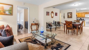 A spacious living area with a glass coffee table, a sofa, and a dining table with chairs. There is a kitchen in the background with wooden cabinets. - 5BR Family Oasis w/ Pool, Near Beach & Downtown (Hollywood)