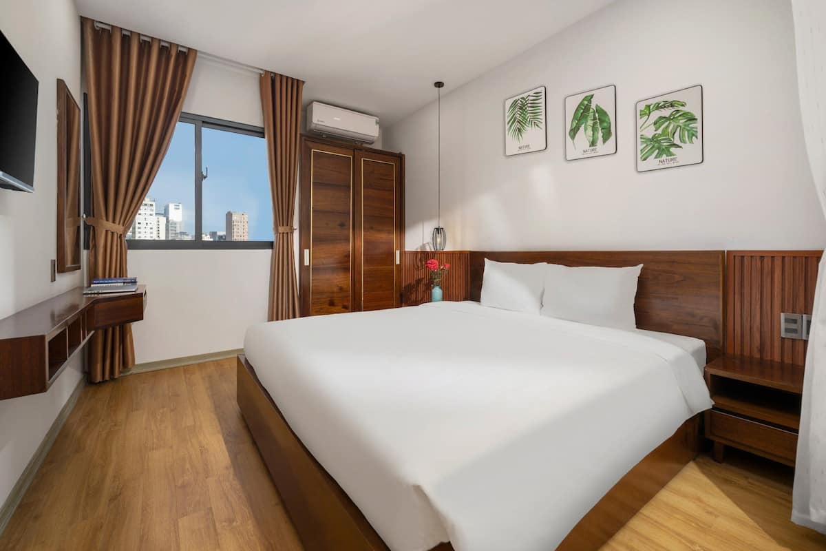 Ein Hotelzimmer mit einem großen Bett, einem Holz-Kopfende, einem Kleiderschrank, einem Schreibtisch mit Stuhl und einem Fenster mit Vorhängen. Über dem Bett befinden sich drei Wandbilder mit Naturmotiven.