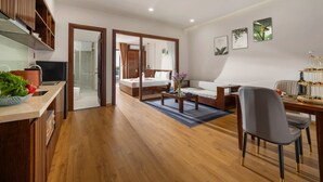 Una habitación de hotel moderna con piso de madera, un área de comedor con mesa y sillas, un sofá y una cama con puerta de vidrio hacia el baño.