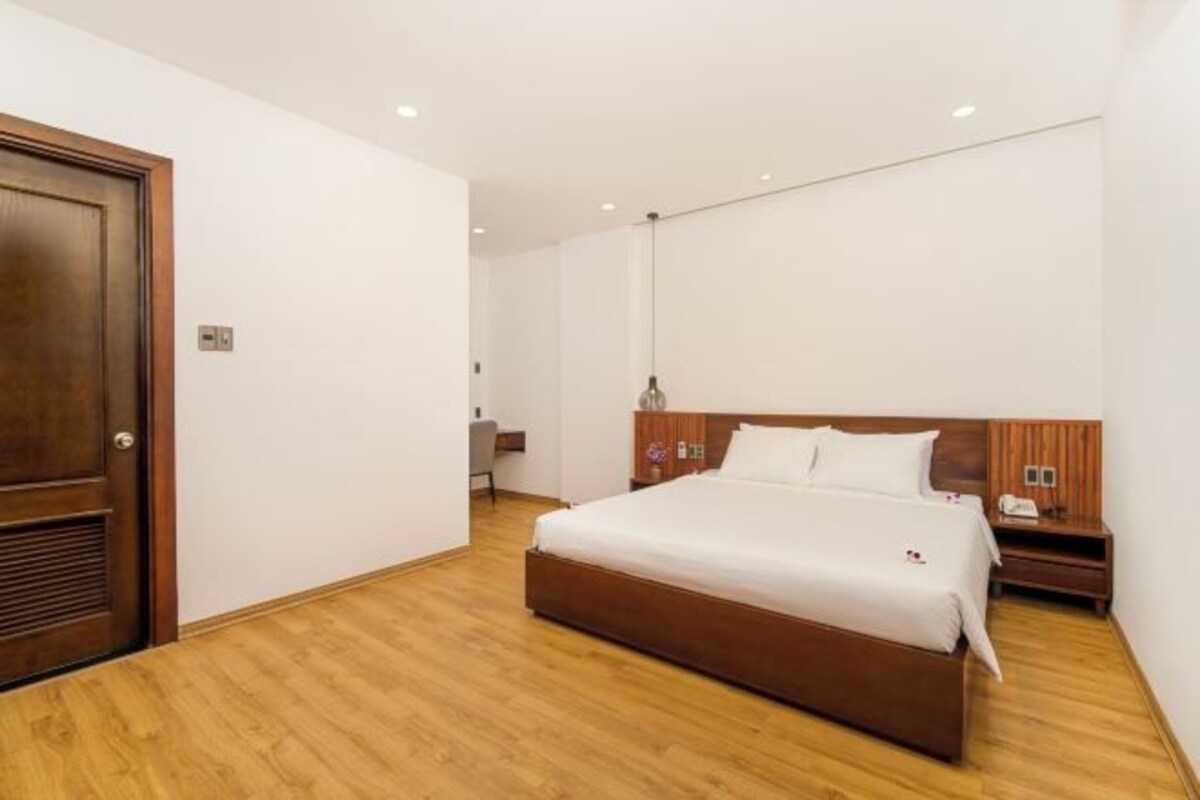 Una habitación de hotel con piso de madera, una cama grande con ropa blanca, cabecera de madera, una mesita de noche con lámpara y una puerta de madera.