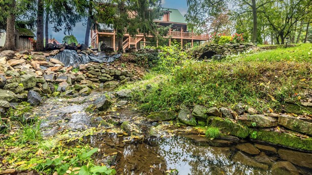 Una cabaña rústica con un puente de piedra sobre un arroyo, rodeada de exuberante vegetación y árboles.
