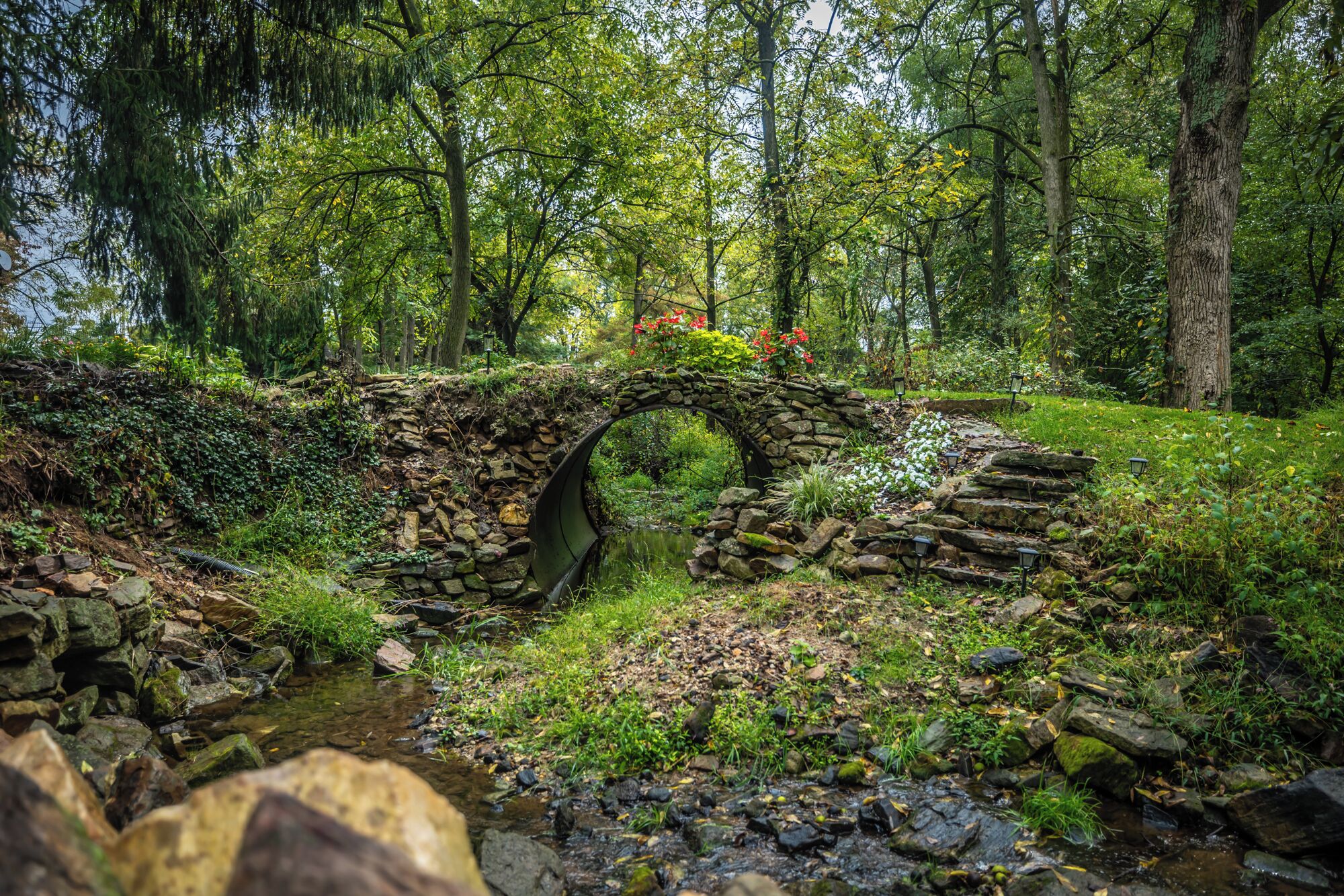 A stone bridge over a stream in a forested area with lush greenery and trees.