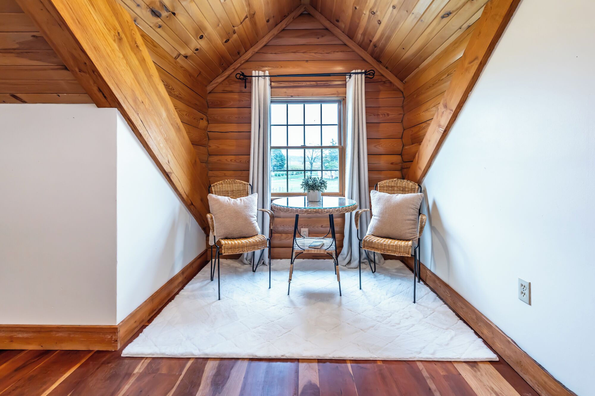 Ein gemütliches Dachzimmer mit Holzwänden, einem großen Fenster, zwei Korbstühlen, einem kleinen Tisch und einem weißen Teppich.