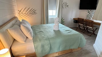 A neatly made bed with a green quilt, a wooden headboard, and a bedside lamp. A wooden desk with two chairs is against the wall, and a flat-screen TV is mounted above it.