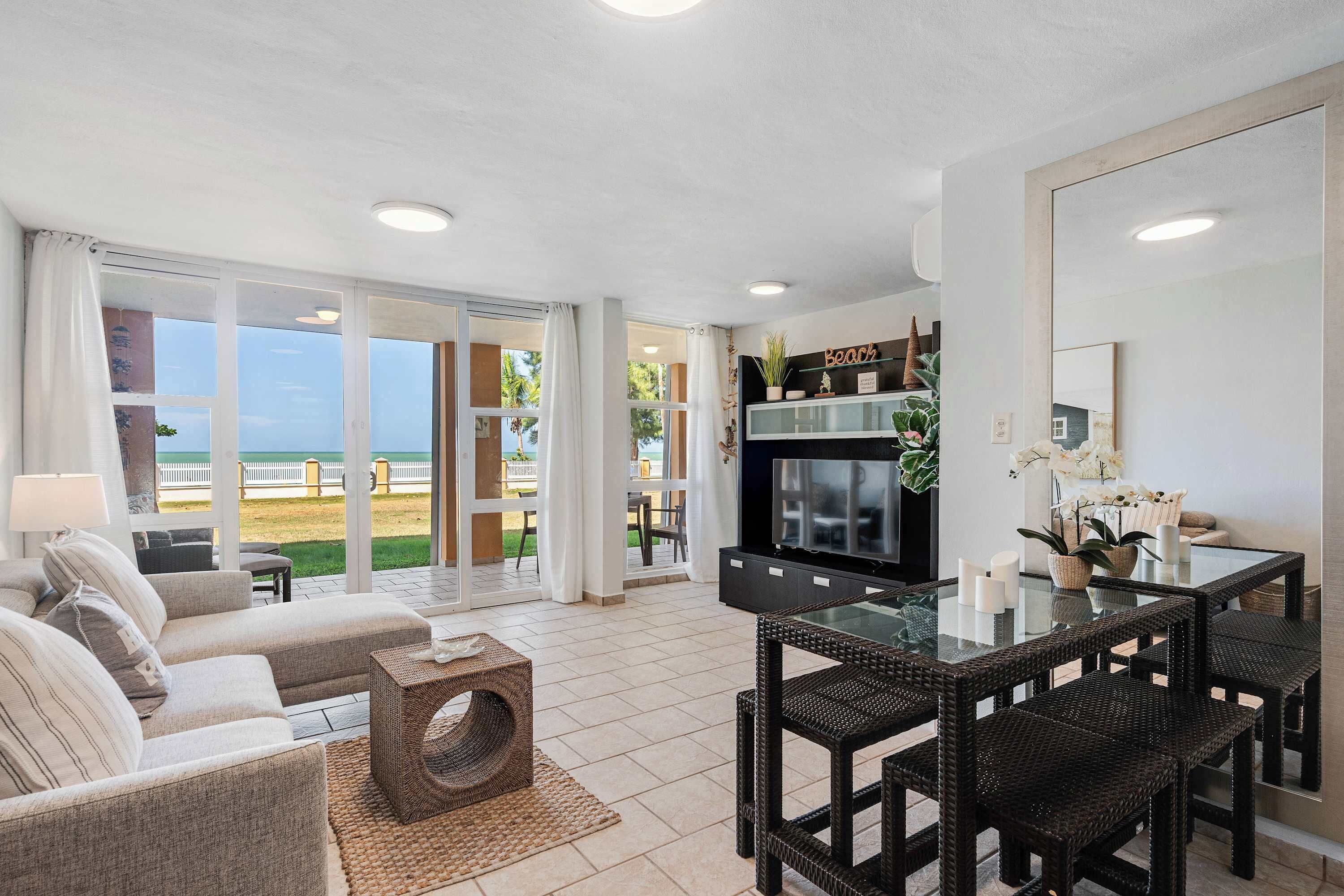 A spacious living area with a sofa set, a glass-top dining table with wicker chairs, a flat-screen TV, and a kitchen island with plants.