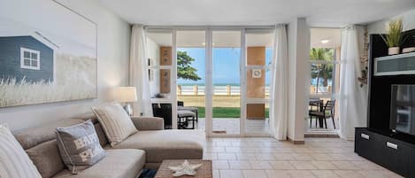 A living room with a sofa, a coffee table, and a TV. There is a sliding glass door leading to an outdoor area with a view of the beach.