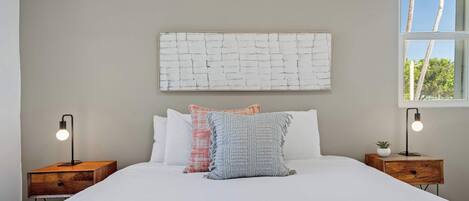 Une chambre à coucher avec un grand lit, deux tables de chevet, un tableau abstrait accroché au mur et une fenêtre donnant sur des arbres.