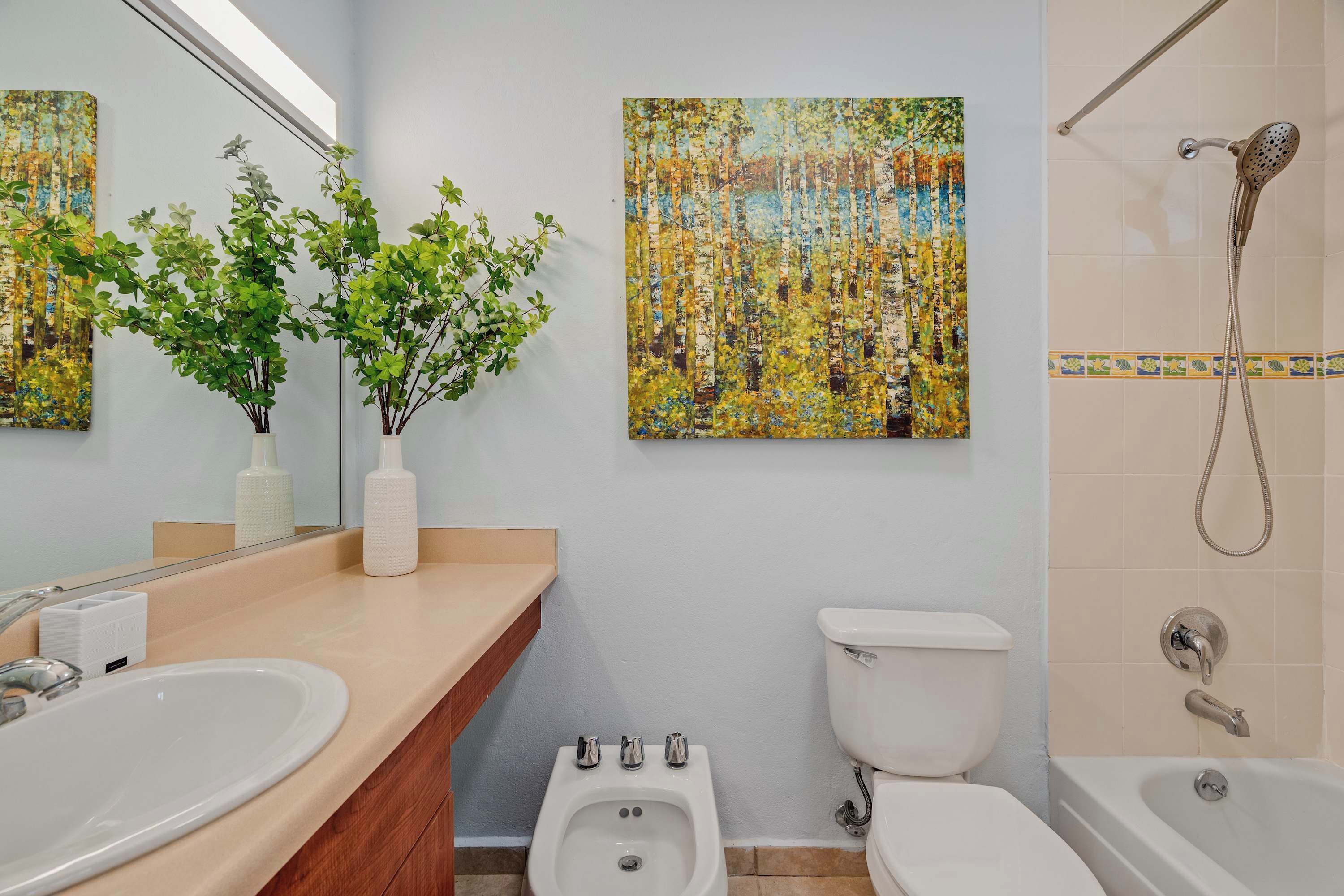 A modern bathroom with a sink, bidet, toilet, and bathtub. There are two wall-mounted paintings and a potted plant on the vanity.
