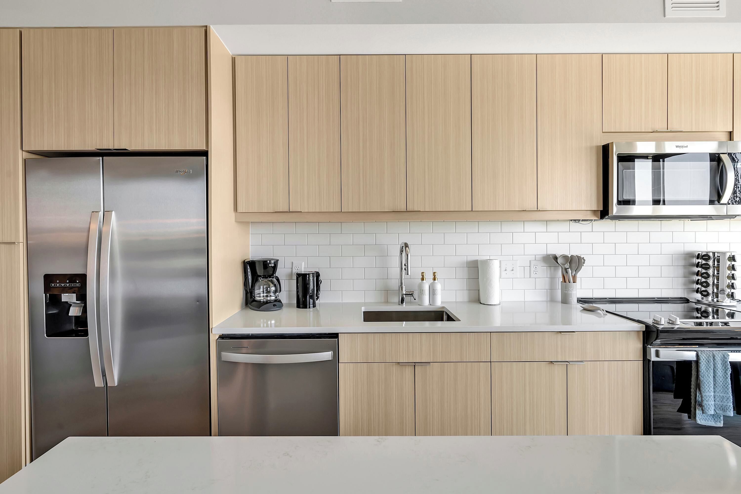 A modern kitchen with light wood cabinets, stainless steel appliances, a white countertop, and a tiled backsplash.