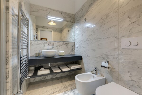 A modern bathroom with a marble floor, a large mirror, a white sink, a bidet, and a towel rack.
