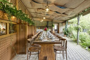 A long wooden table set with plates, cups, and a vase of flowers, surrounded by chairs in a covered outdoor dining area with plants and a stone wall.