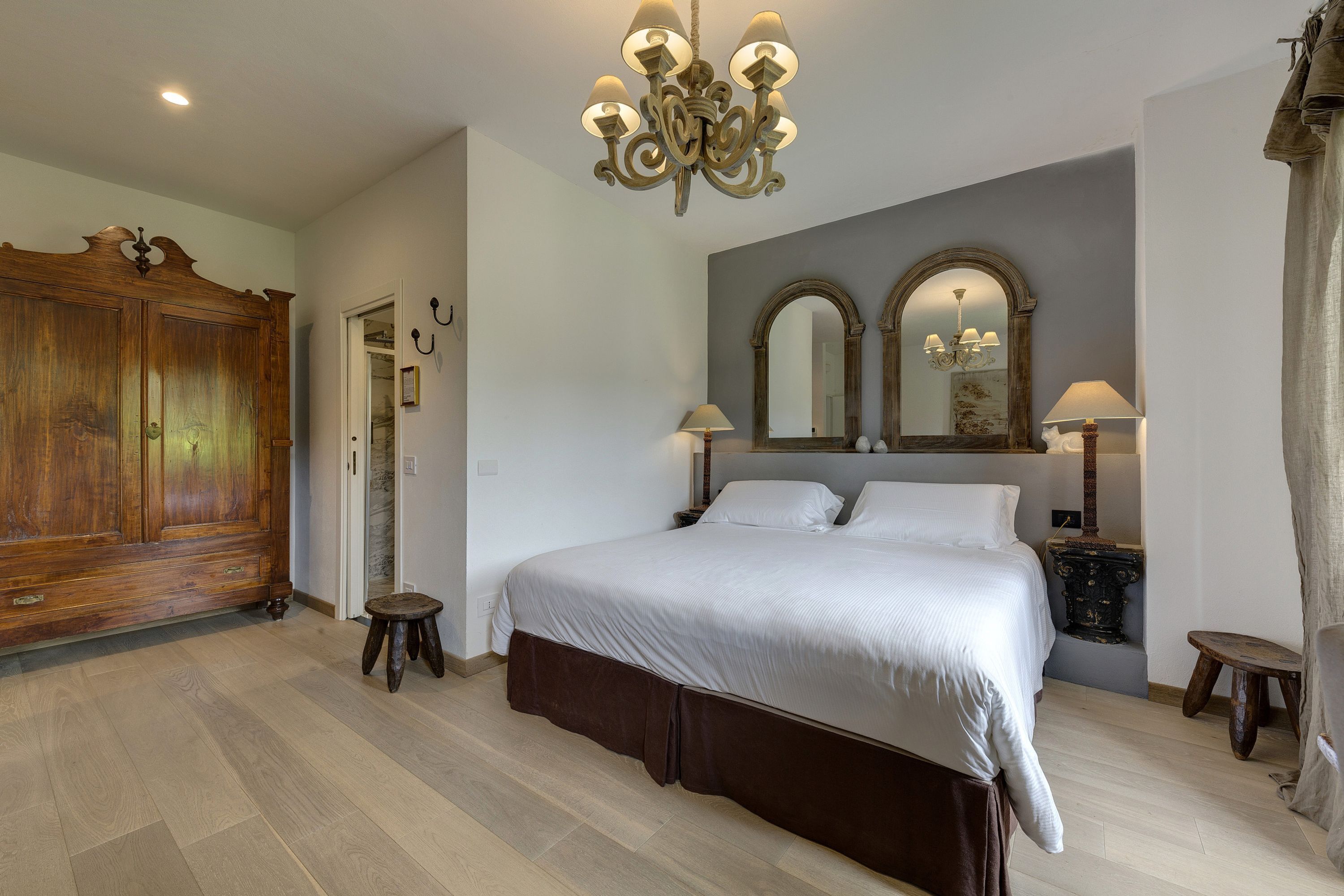 A bedroom with a large bed, a wooden wardrobe, a chandelier, and two wall-mounted mirrors.