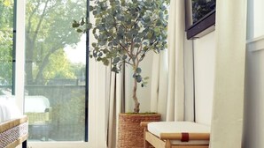 A room with a large window, a wooden table, a wicker chair, a blue rug, and a potted plant. - Austin Abode | Heated Pool | Sleeps 12 | Rainey St (Austin)