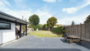 A patio area with a stone floor, a wooden table, and a lawn. - Charming 3-Bedroom Bungalow in London (Greater London)