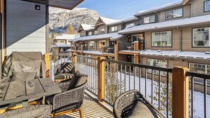Un espacio de descanso techado al aire libre con sillas y una parrilla, con vistas a un conjunto de edificios cubiertos de nieve y con numerosas ventanas, y un paisaje montañoso de fondo.