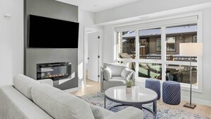 Una sala de estar moderna con televisor de pantalla plana, chimenea, sofá gris, una mesa redonda y ventanas amplias con vistas al exterior.
