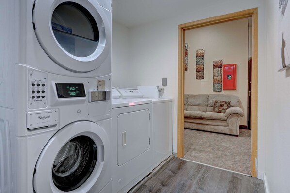 A laundry room with a front-loading washer and dryer, a small table with a whiteboard, and a beige sofa in the adjacent living area.