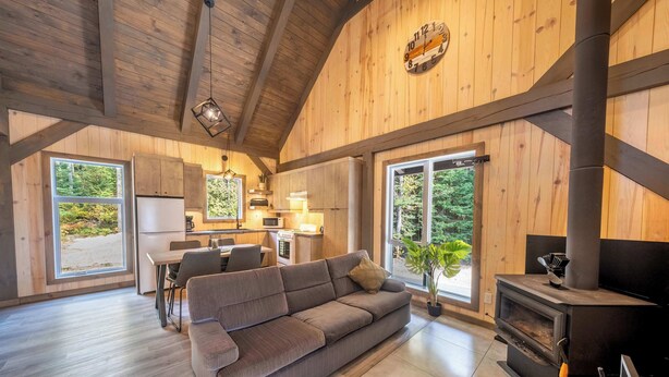 A spacious living area with a sofa, a fireplace, and a kitchen in the background. - Spa and Nature | Chalet des Pins Rouge (Sainte-Euphémie-sur-Rivière-Sud)