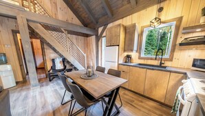 A wooden interior with a dining table, chairs, a kitchen with a window, and a staircase. - Spa and Nature | Chalet des Pins Rouge (Sainte-Euphémie-sur-Rivière-Sud)