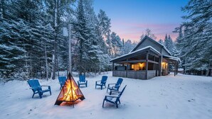 A lodge with a wooden exterior, surrounded by snow-covered trees, and a fire pit with chairs in the foreground. - Spa and Nature | Chalet des Pins Rouge (Sainte-Euphémie-sur-Rivière-Sud)