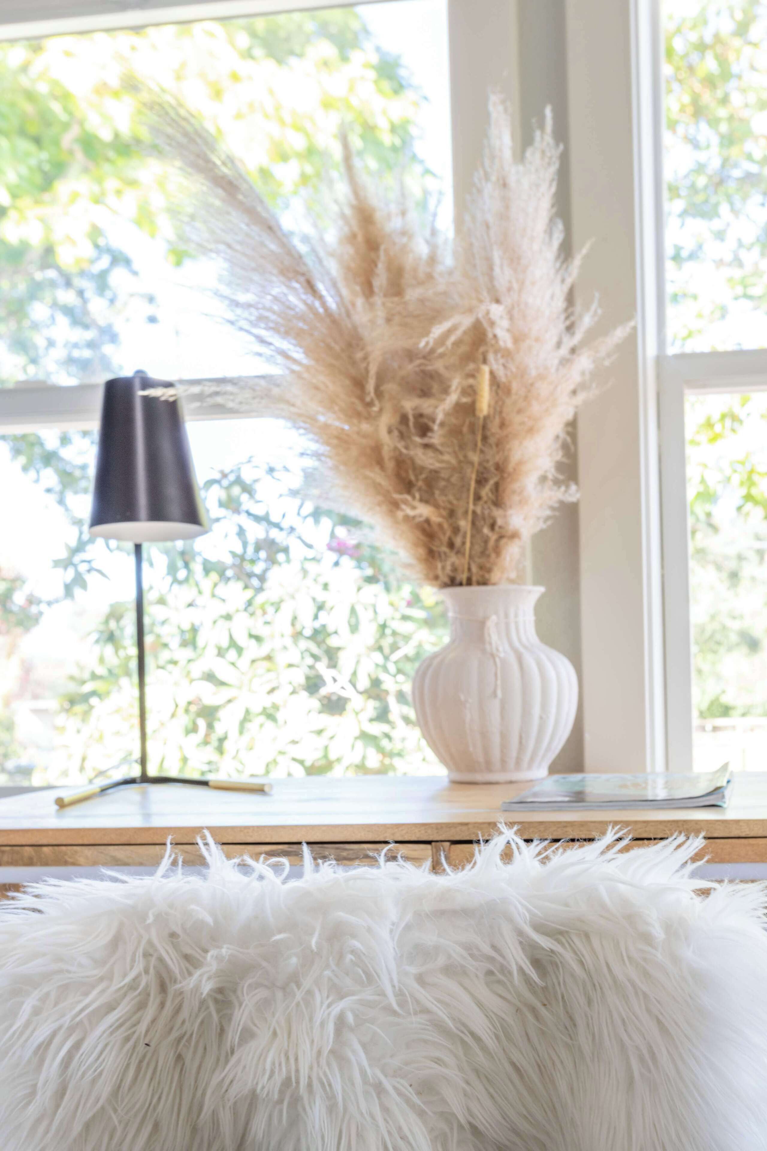 A white vase with pampas grass on a wooden table near a window.