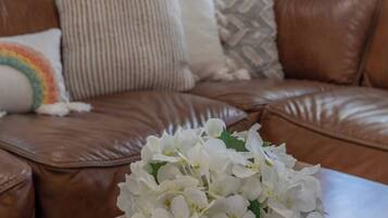 Ein braunes Ledersofa mit gemusterten Kissen, ein runder Spiegel an der Wand und eine Vase mit weißen Blumen auf einem Holztisch.
